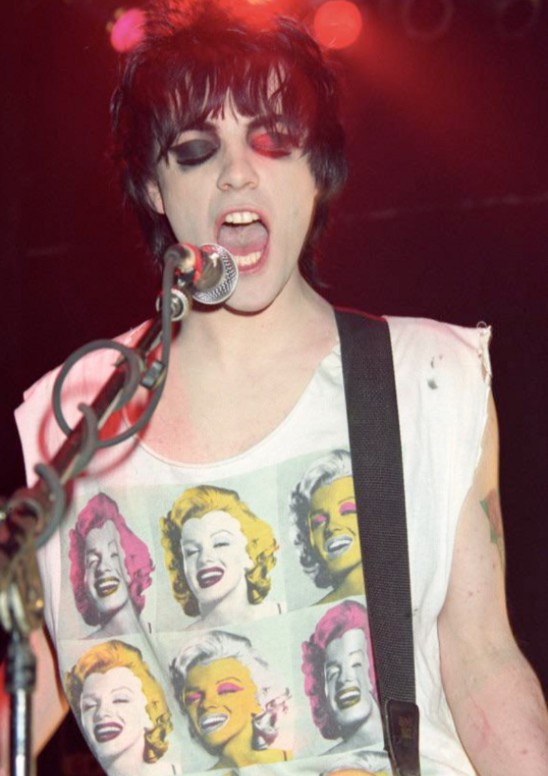 A musician with dramatic black and red eye makeup sings into a microphone while playing guitar. They wear a sleeveless shirt featuring colorful pop art images of Marilyn Monroe. Red stage lights glow in the background.