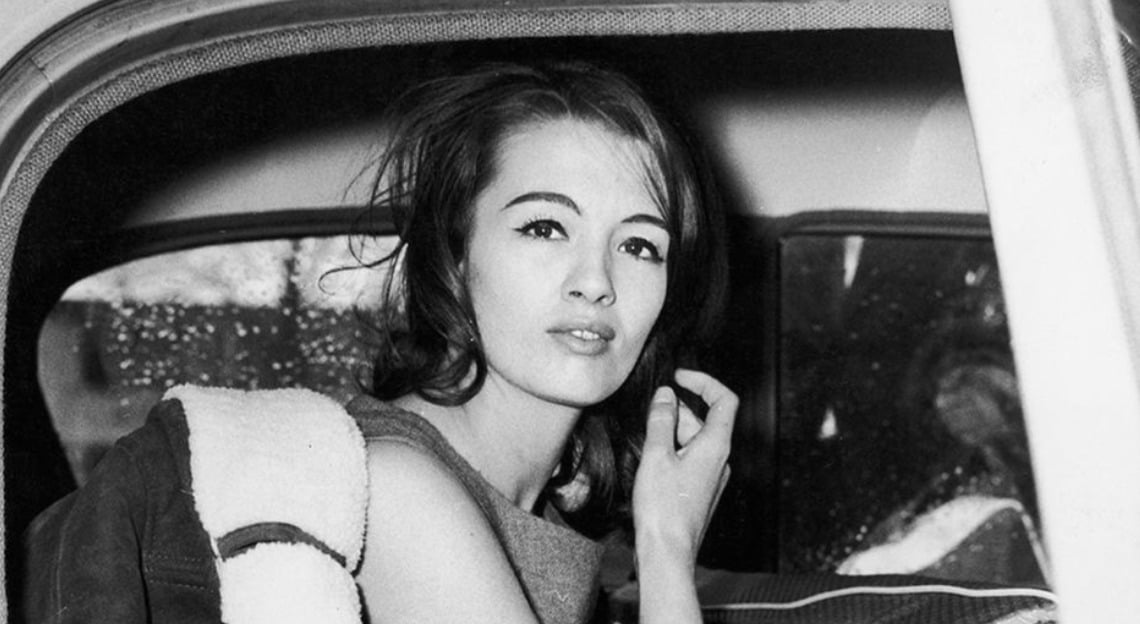 A young woman with long dark hair sits in the backseat of a car, looking out the open window with a thoughtful expression. She wears a sleeveless top and holds her hand near her face. The window shows raindrops outside.