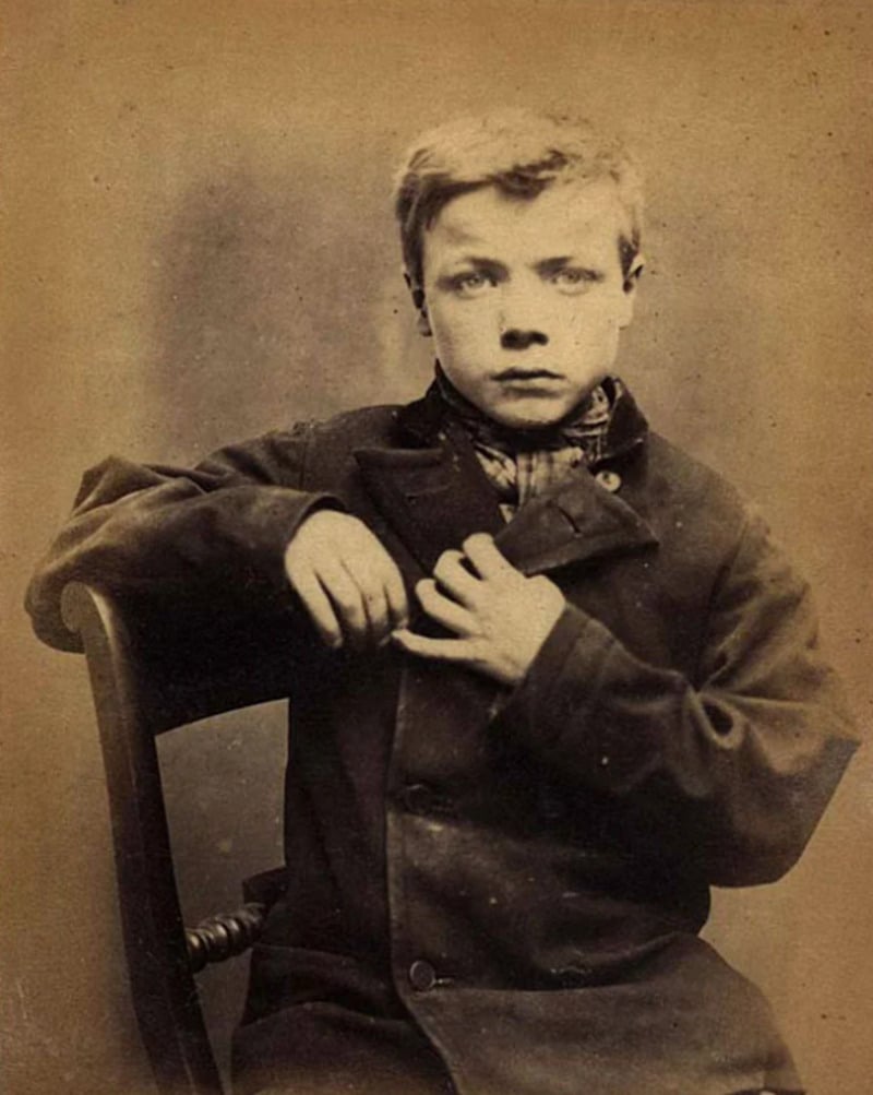 A sepia-toned vintage photograph of a young boy with light hair, wearing a dark coat and plaid scarf, sitting on a wooden chair and looking seriously at the camera.