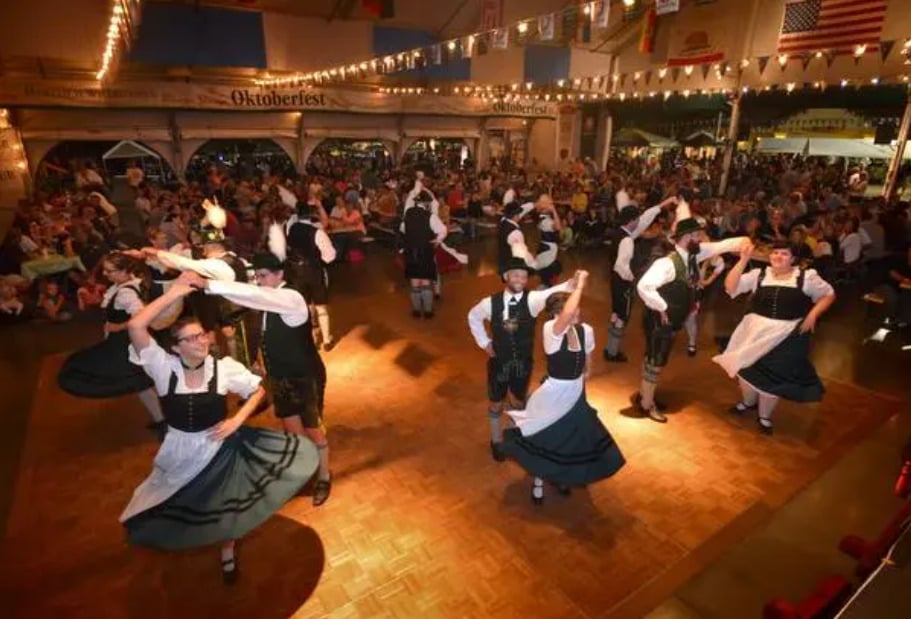 A group of men and women in traditional Bavarian clothing perform a folk dance on a wooden floor at an Oktoberfest celebration, with a large audience seated around them under festive string lights.