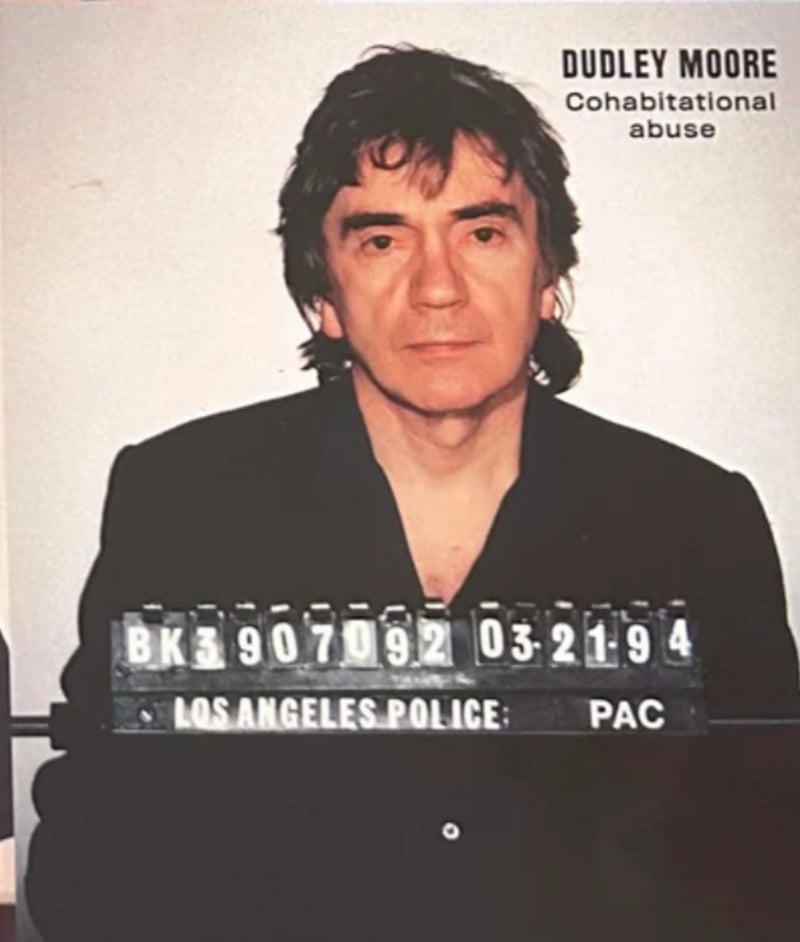 A man with dark hair wearing a black shirt sits for a mugshot. The sign reads "LOS ANGELES POLICE" with booking numbers. Text in the upper right corner says "DUDLEY MOORE Cohabitational abuse.