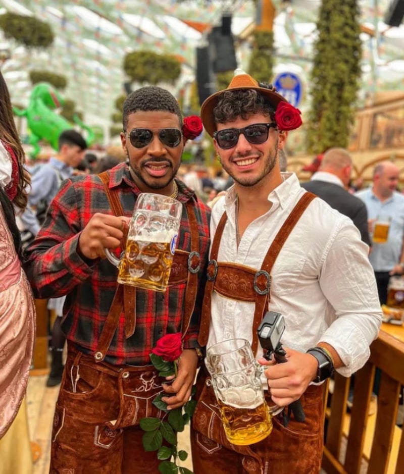 Two men smile at a festival, each holding a large beer mug and a red rose. They wear traditional Bavarian lederhosen; one sports a checkered shirt, the other a white shirt with sunglasses. The background shows a busy, festive crowd.