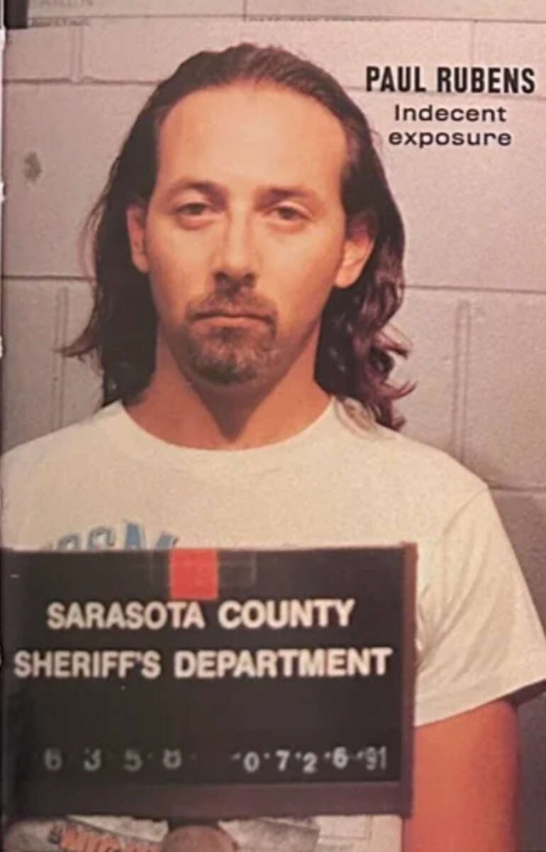 A man with long hair and a goatee is shown in a mugshot holding a SARASOTA COUNTY SHERIFF'S DEPARTMENT sign. Text beside him reads "Paul Rubens Indecent exposure.