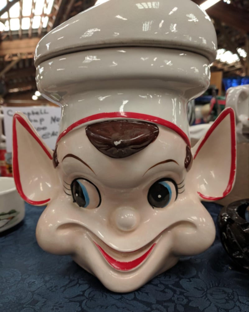 A ceramic cookie jar shaped like an elf's head with large ears, a mischievous smile, blue eyes, and a white chef's hat, displayed on a blue tablecloth at an indoor market.