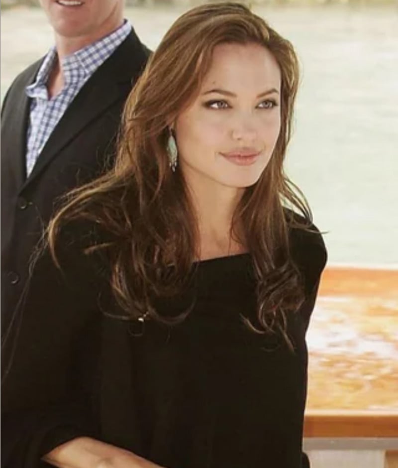 A woman with long brown hair and light eyes, wearing a black top and earrings, stands outdoors near water. Another person in a suit is partially visible beside her.