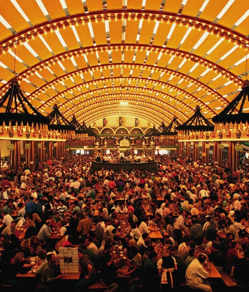 A large, crowded beer hall with rows of long wooden tables filled with people. The arched ceiling is lit with bright yellow lights, and a stage with musicians is visible at the far end. The atmosphere is lively and festive.