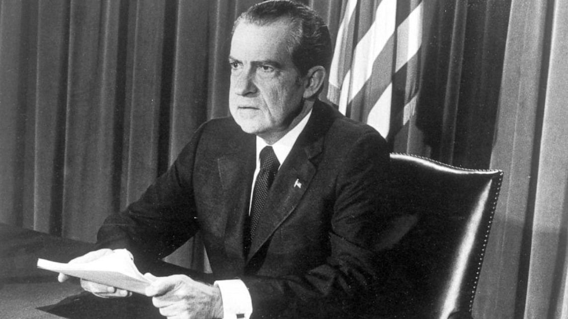 A man in a suit sits at a desk holding papers, with an American flag and curtains in the background. He has a serious expression and appears to be addressing an audience or giving a speech.
