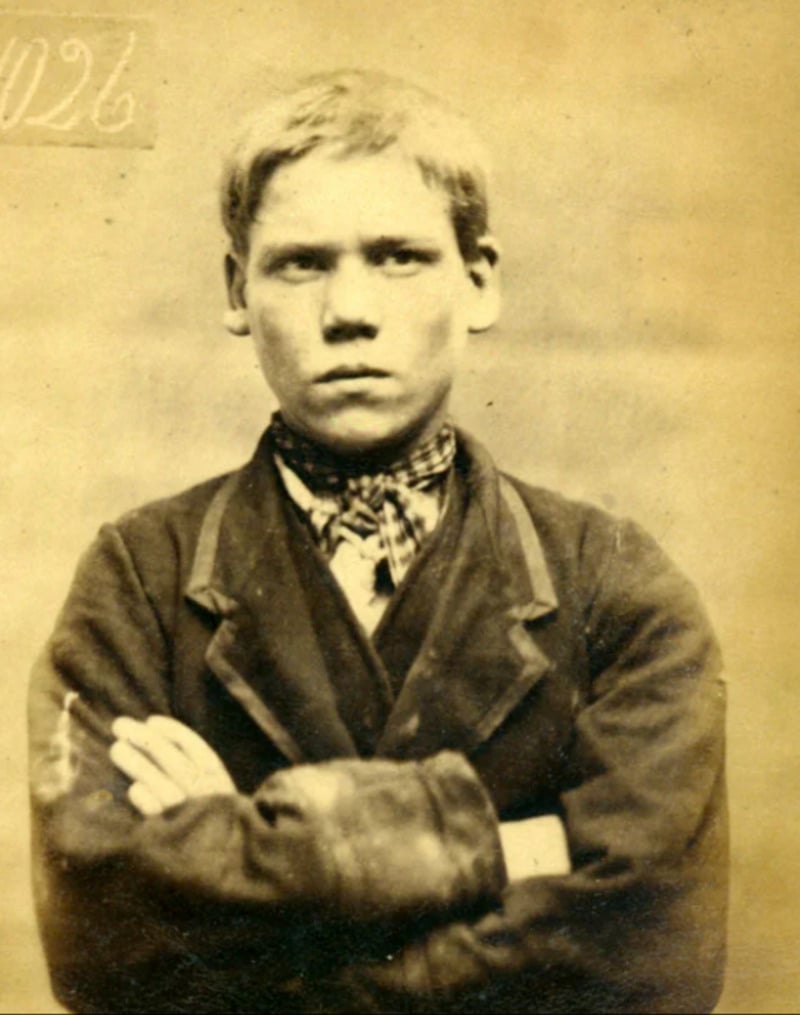 Sepia-toned portrait of a young boy wearing a suit jacket and patterned scarf, with his arms crossed and a serious expression, against a plain background. A faint handwritten number appears in the top left corner.