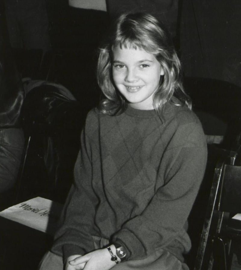 A young girl with shoulder-length hair sits and smiles at the camera. She is wearing a long-sleeve, diamond-patterned sweater and light pants. The photo is black and white, with empty chairs in the background.