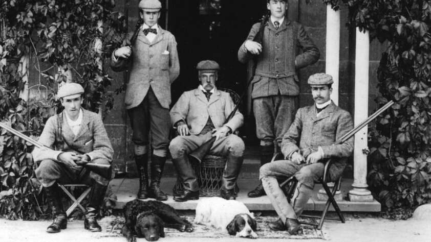 Five men in vintage hunting attire pose with rifles outside a building, with two dogs lying at their feet. Three men are seated, while two stand behind them. The setting is leafy and the photo is in black and white.