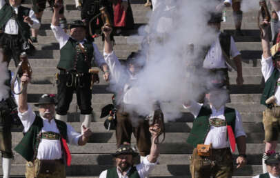 A group of men in traditional Bavarian clothing stand on stone steps, raising rifles and firing them, with smoke billowing around. They wear hats, vests, and leather shorts, participating in a festive event.