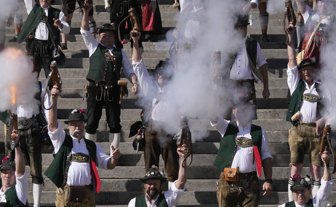 A group of men in traditional Bavarian clothing stand on steps, raising rifles or hand cannons, with smoke billowing from some of the guns, as part of a ceremonial or festive event.