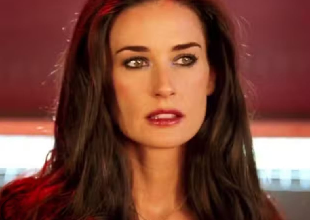 A woman with long dark hair and striking green eyes looks slightly to the side. She has light makeup with bold eyeliner and a neutral lipstick. The background is softly lit with red tones.