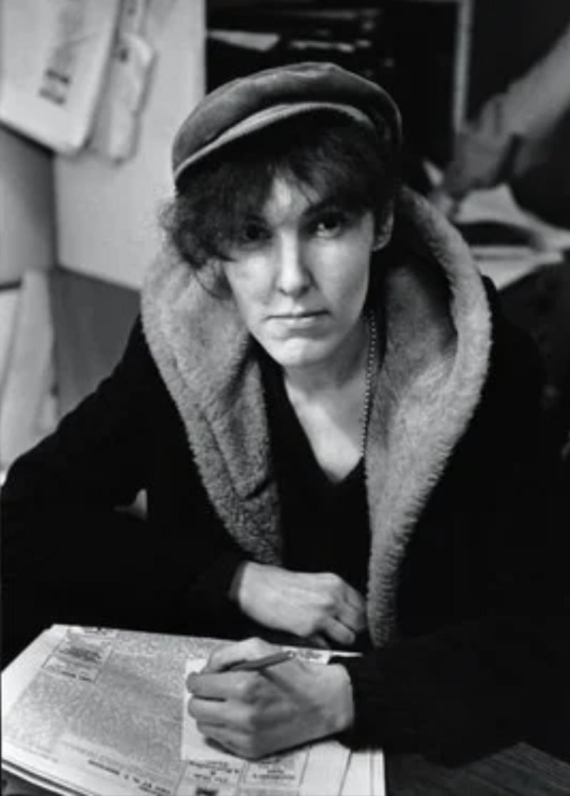 A person wearing a cap and a coat with a fur collar sits at a desk, looking at the camera while holding a pencil and writing on newspaper pages. Papers and folders are visible in the background.