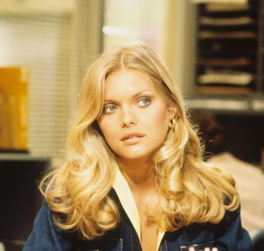 A woman with long, wavy blonde hair and blue eyes sits indoors, looking slightly to the side. She wears a denim jacket and gold hoop earrings. Office shelves and files are visible in the background.
