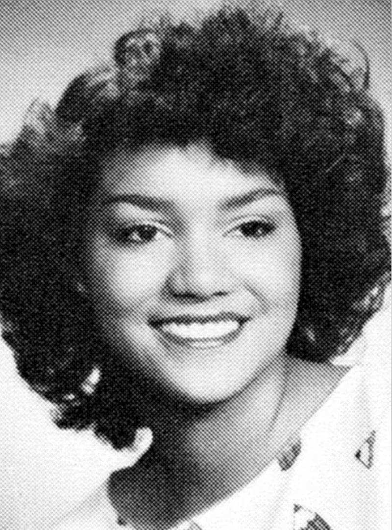 Black and white portrait of a young woman with curly hair, smiling and facing slightly to the side. She is wearing a light-colored top with a subtle pattern. The background is plain.