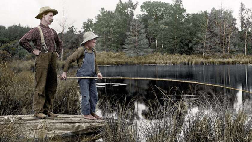 Two people stand on a wooden raft near the edge of a pond, surrounded by tall grass and trees. One holds a fishing rod while the other watches, both dressed in old-fashioned clothes and hats.