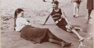 A woman in a dress and boots sits on the sand beside a man in a &ldquo;Swimming Instructor&rdquo; tank top and shorts, relaxing at the beach. Other people walk nearby along the shoreline. The image is in black and white.