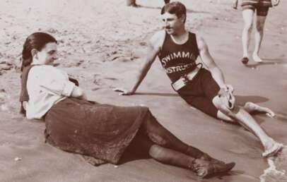 A woman in a dress and boots sits on the sand beside a man in a “Swimming Instructor” tank top and shorts, relaxing at the beach. Other people walk nearby along the shoreline. The image is in black and white.
