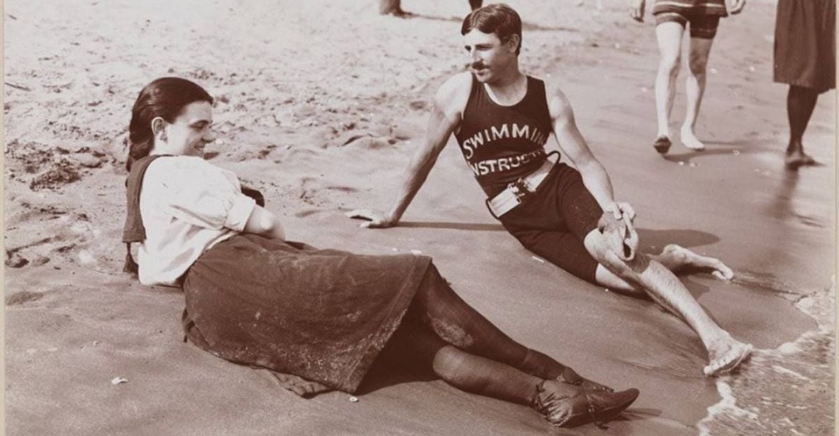 A woman in a dress and boots sits on the sand beside a man in a “Swimming Instructor” tank top and shorts, relaxing at the beach. Other people walk nearby along the shoreline. The image is in black and white.