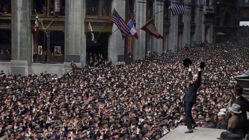 A massive crowd fills a city street, cheering as a man on a platform waves his hat. Several flags, including the American, French, and Italian flags, hang from the building above the crowd.