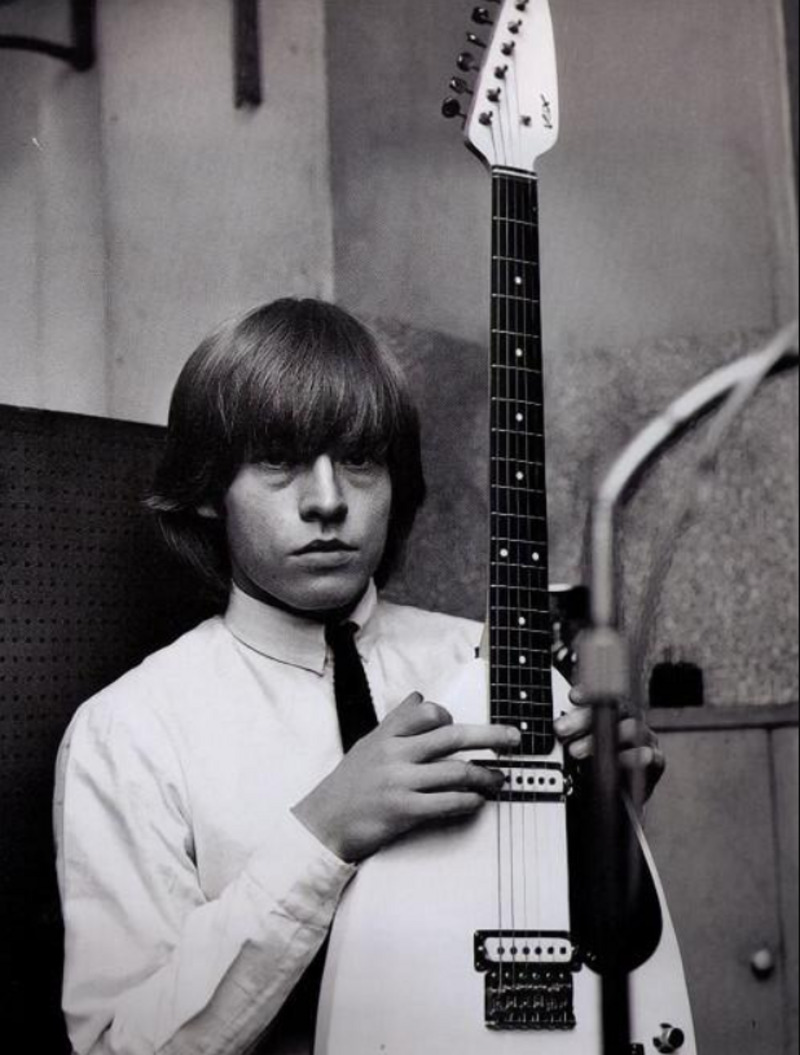 A young man with medium-length hair and a serious expression holds an electric guitar vertically in a recording studio. He is wearing a light-colored shirt and a dark tie.