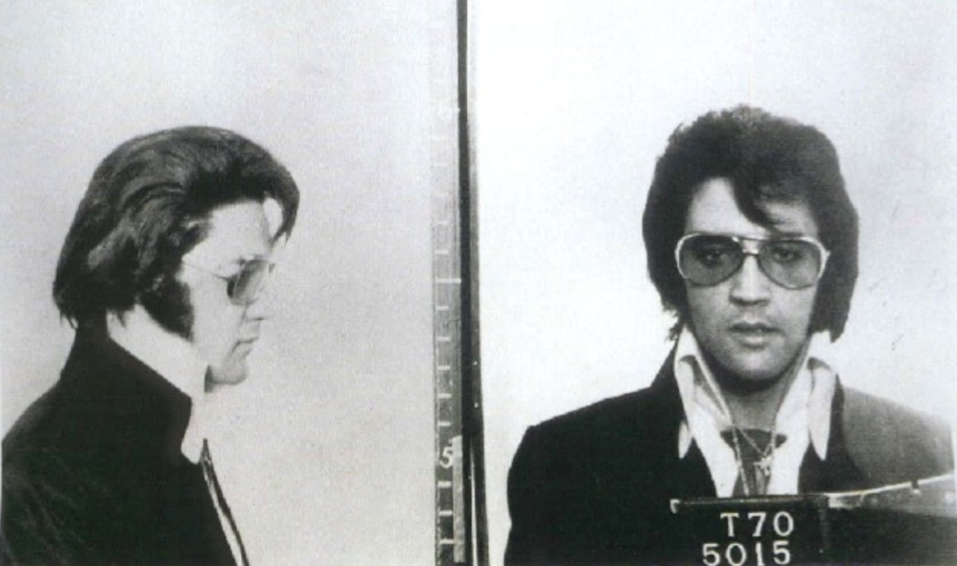Black and white mugshot of a man with dark hair and sunglasses shown from the side and front wearing a collared shirt and jacket, holding a placard with identification numbers.