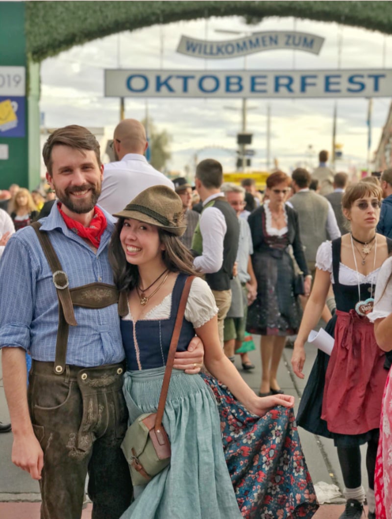 A smiling couple in traditional Bavarian outfits poses in front of a crowd at Oktoberfest, with a large “Willkommen zum Oktoberfest” sign overhead. The woman holds her dress while the man stands beside her.