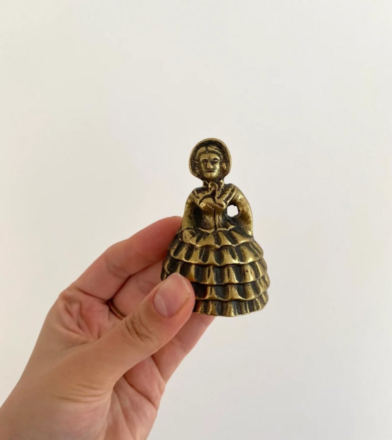 A hand holding a small brass figurine of a woman in a ruffled dress with a bonnet, against a plain white background.