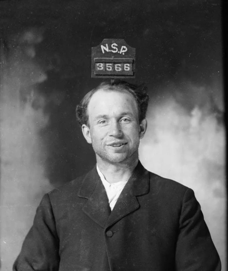 Black-and-white photo of a man in a suit, smiling, with an identification board above his head displaying "N.S.P. 3556" against a plain studio backdrop.