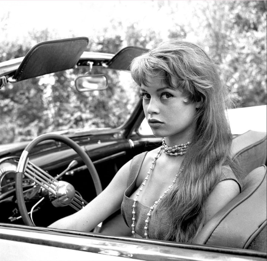 A young woman with long hair and a pearl necklace sits in the driver’s seat of a classic convertible car, looking toward the camera. The photo is black and white, and trees are visible in the blurred background.