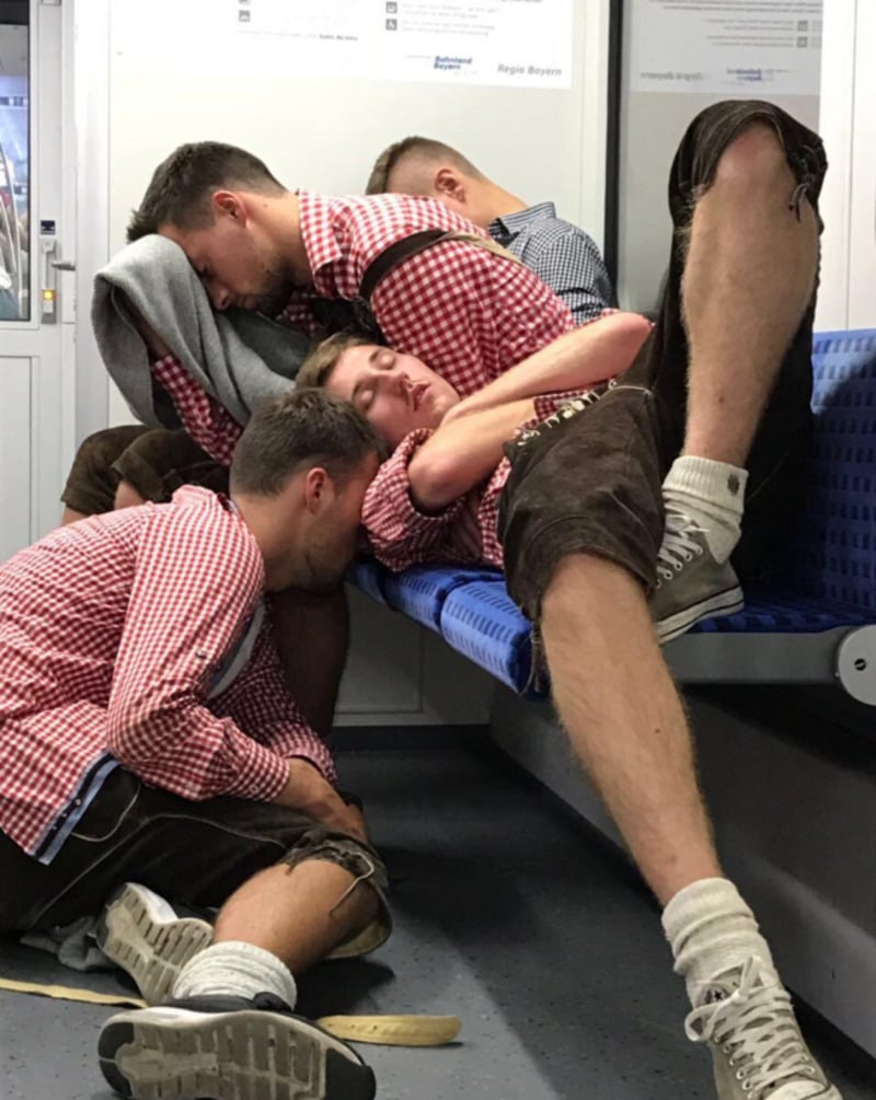 Four young men in red checkered shirts and shorts are sleeping close together on blue seats in a train, with some sitting and others laying across the bench and floor.