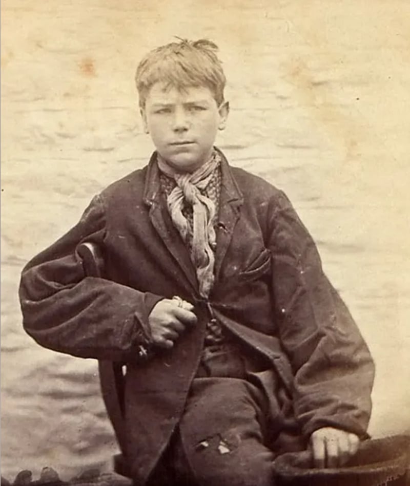 A young boy with short hair sits facing the camera, wearing a worn jacket, neckerchief, and pants with holes. His expression is serious, and he holds a hat in his lap. The background is plain and light-colored.