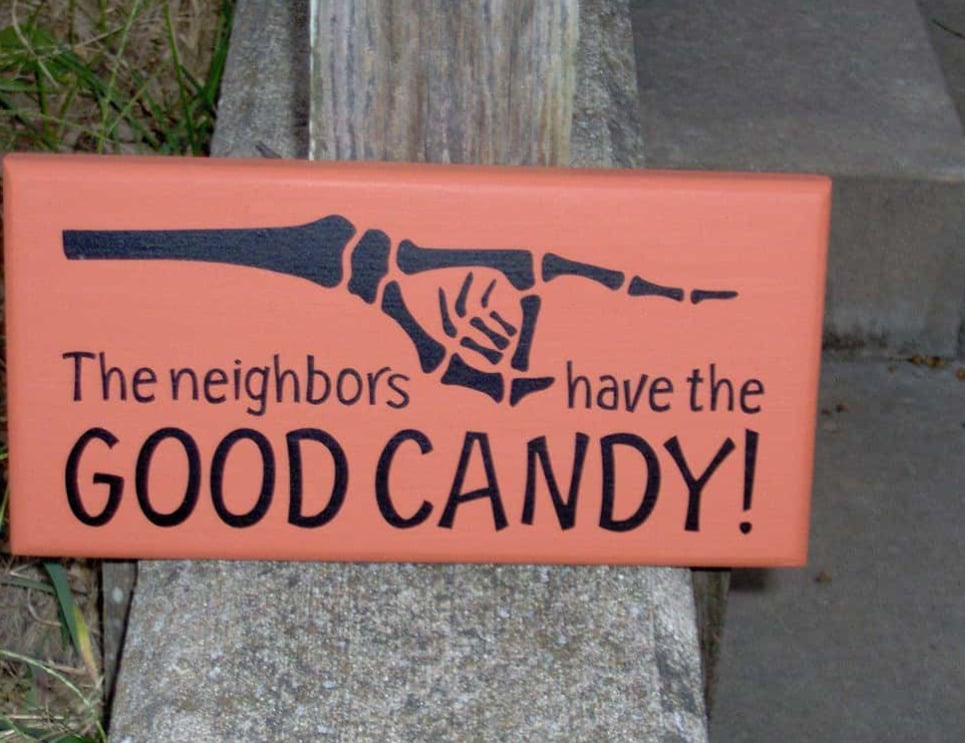 A Halloween sign with a skeleton hand pointing right reads: "The neighbors have the GOOD CANDY!" The sign is propped up outdoors by some steps.