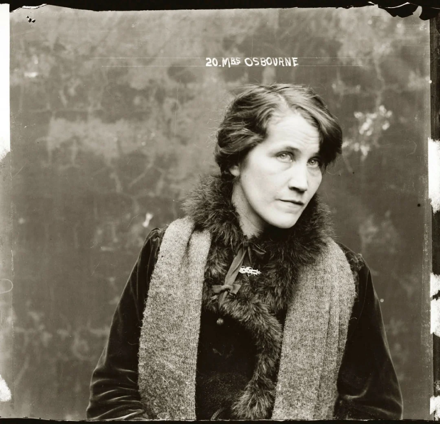 A black-and-white portrait of a woman with braided hair, wearing a dark dress and a fur-collared scarf. She looks slightly to the side with a serious expression. Text above her reads “20. MRS OSBOURNE.”