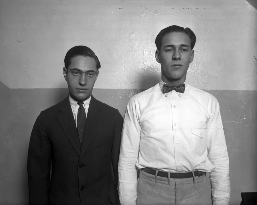 Two young men stand side by side in front of a plain, two-toned wall. The man on the left wears a dark suit and tie, while the man on the right wears a light shirt with a bow tie and belted trousers.