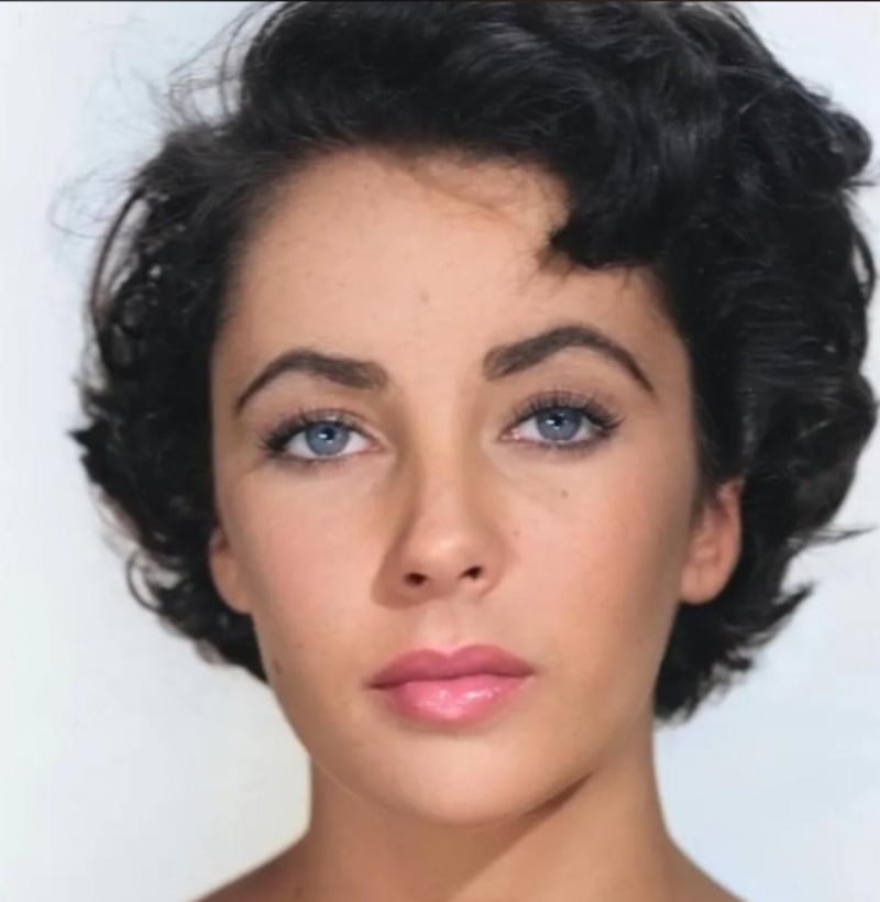A woman with short, dark curly hair, blue eyes, and pink lips poses against a plain light background, looking directly at the camera with a neutral expression.