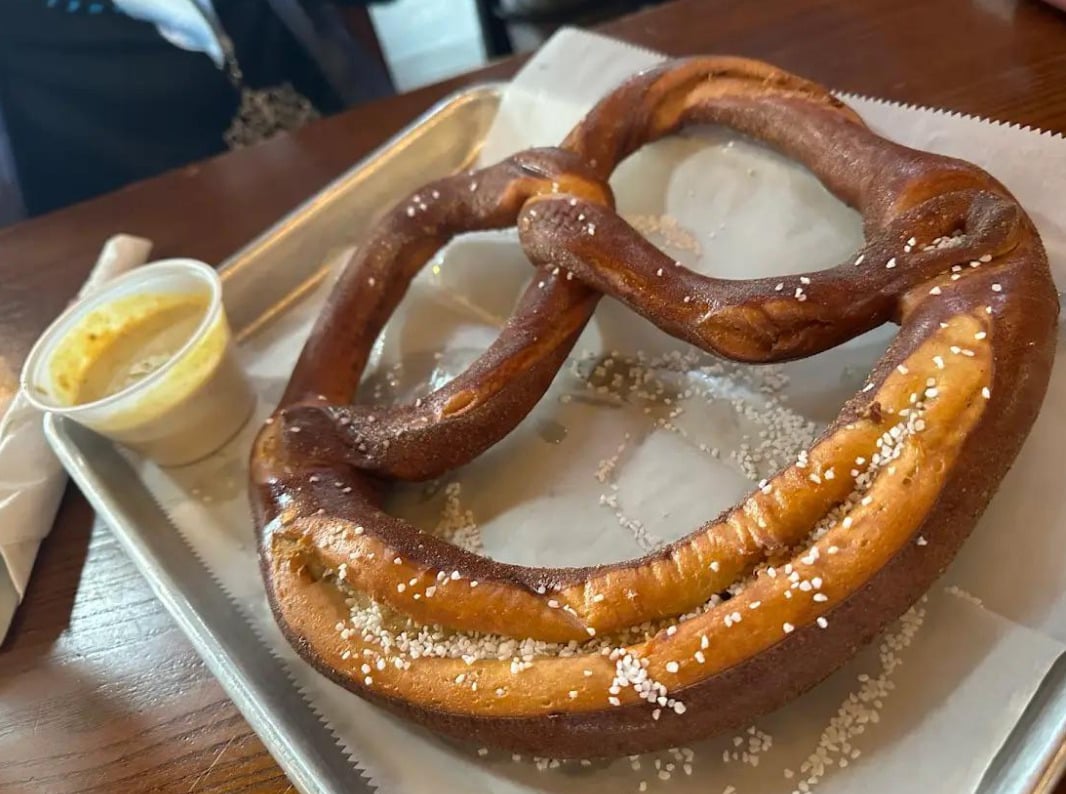 A large soft pretzel topped with coarse salt sits on a metal tray lined with parchment paper, next to a small cup of yellow dipping sauce.