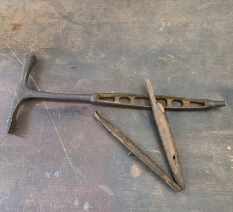 A vintage metal tool with a T-shaped handle and a wooden object split in two, both resting on a worn wooden surface. The tool has holes along its shaft and a tapered end.