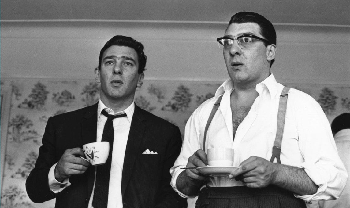 Two men in suits stand indoors, each holding a cup and saucer. One wears glasses and suspenders, the other a dark tie and jacket. Both appear to be looking at something with surprised or intent expressions.