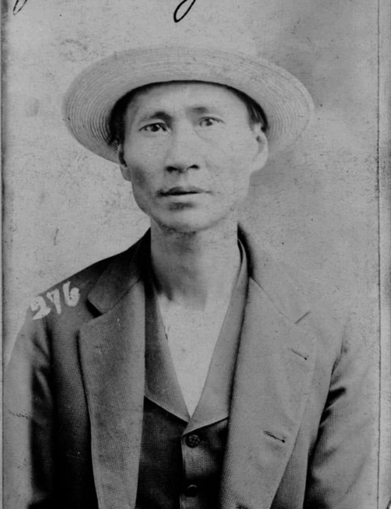 Black and white portrait of a man wearing a light-colored straw hat and a suit jacket with the number 276 on the shoulder, looking directly at the camera with a neutral expression.