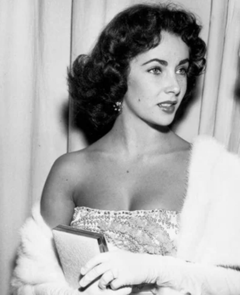 A woman with short, dark curly hair wears a strapless, sequined dress and a white fur stole. She holds a clutch and wears gloves, looking off camera with a composed expression, standing in front of a curtain.