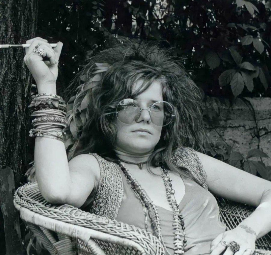 A woman with long wavy hair, round glasses, and layered jewelry relaxes in a wicker chair outdoors, holding a cigarette and looking at the camera. The background features leafy plants and a stone wall.