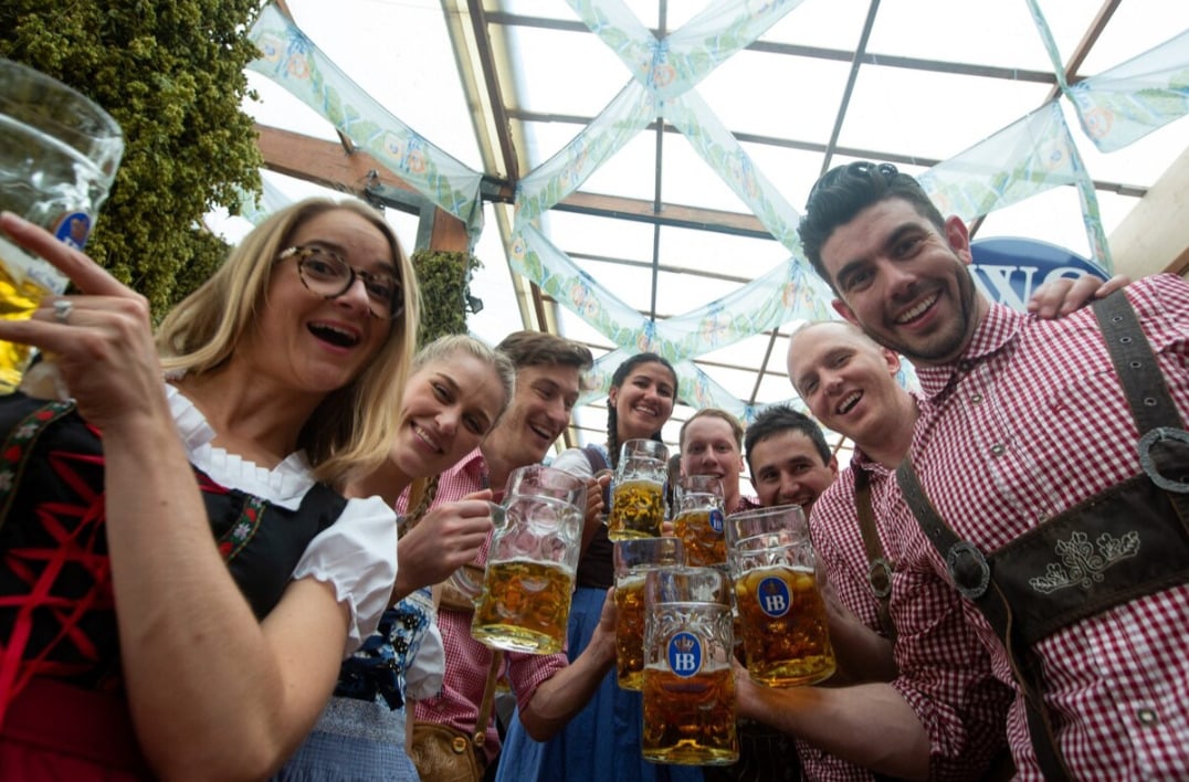 A group of smiling people in traditional Bavarian clothing raise large beer mugs in celebration under a festive, glass-roofed canopy.
