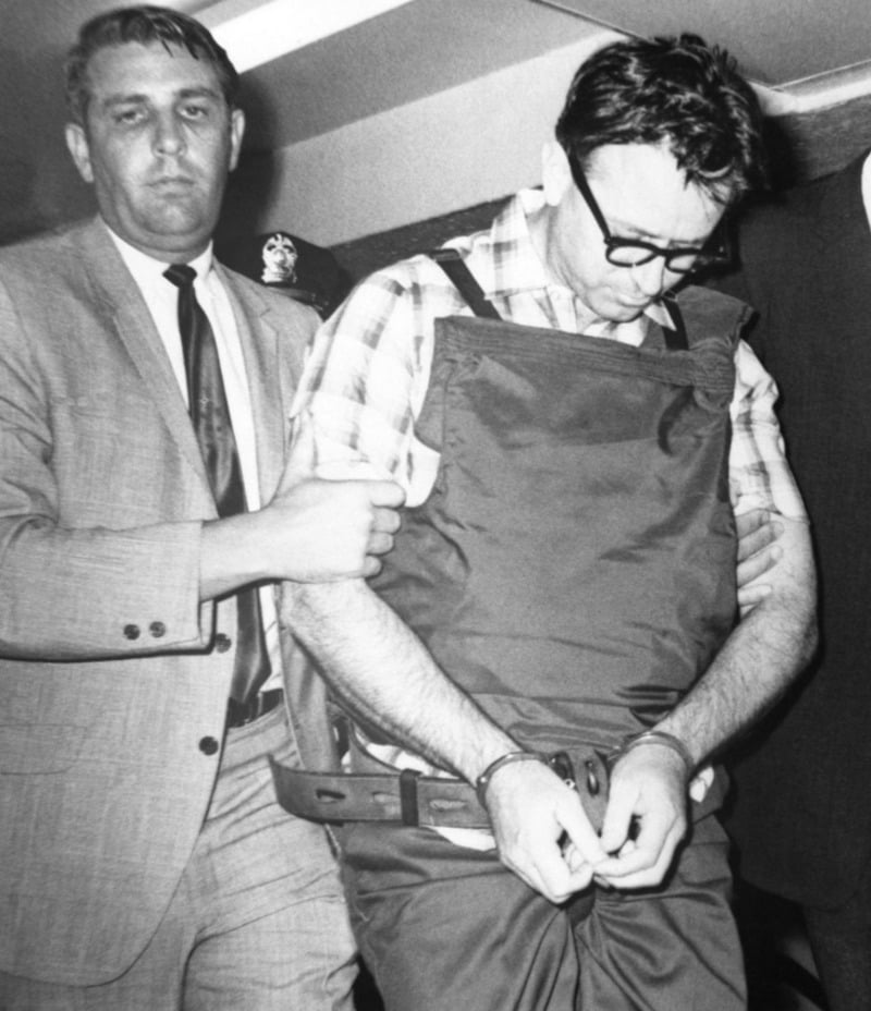 A man in glasses and a bulletproof vest is handcuffed and escorted by another man in a suit and tie. The scene appears serious, possibly relating to a crime or arrest, with multiple people present.