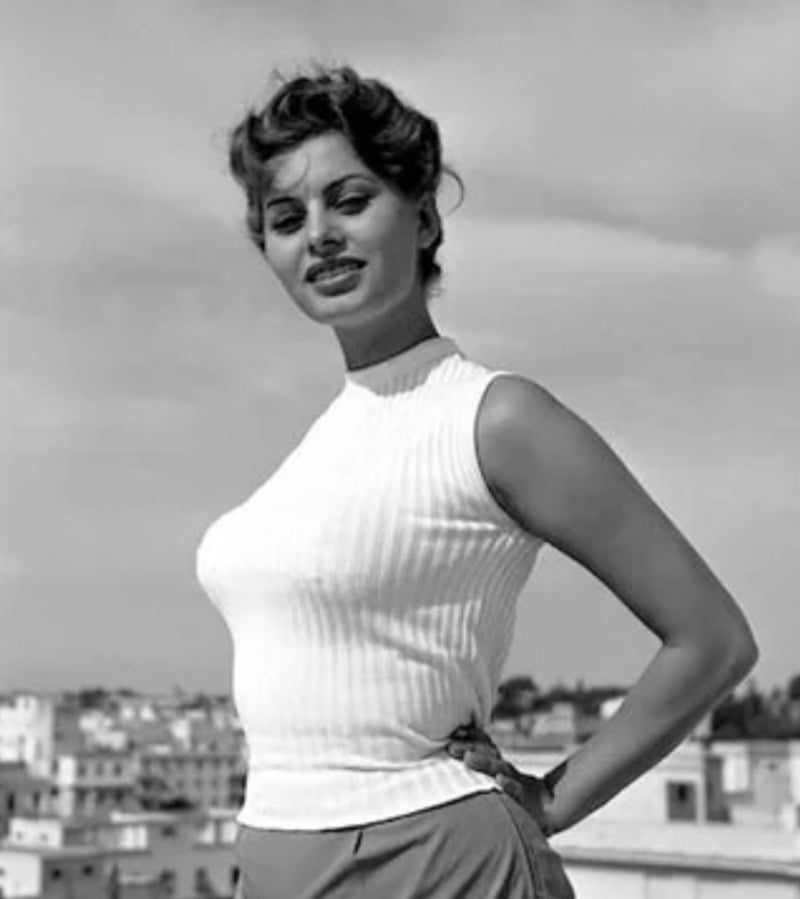 A woman with short curly hair wearing a sleeveless ribbed white top and pants stands outdoors with one hand on her hip, smiling slightly, with buildings and a cloudy sky in the background.
