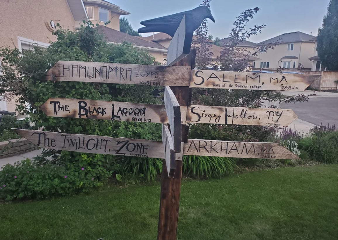 A wooden signpost in a neighborhood yard displays hand-painted arrows pointing to fictional and real locations, including Hamunaptra, Salem, The Black Lagoon, Sleepy Hollow, The Twilight Zone, and Arkham.