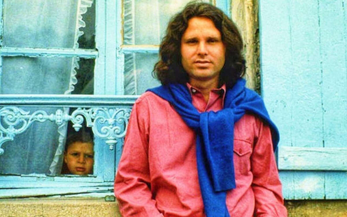 A man with long brown hair, wearing a pink shirt and blue sweater draped over his shoulders, stands in front of a blue window. A child peers through the window behind him, partially hidden by the window frame.
