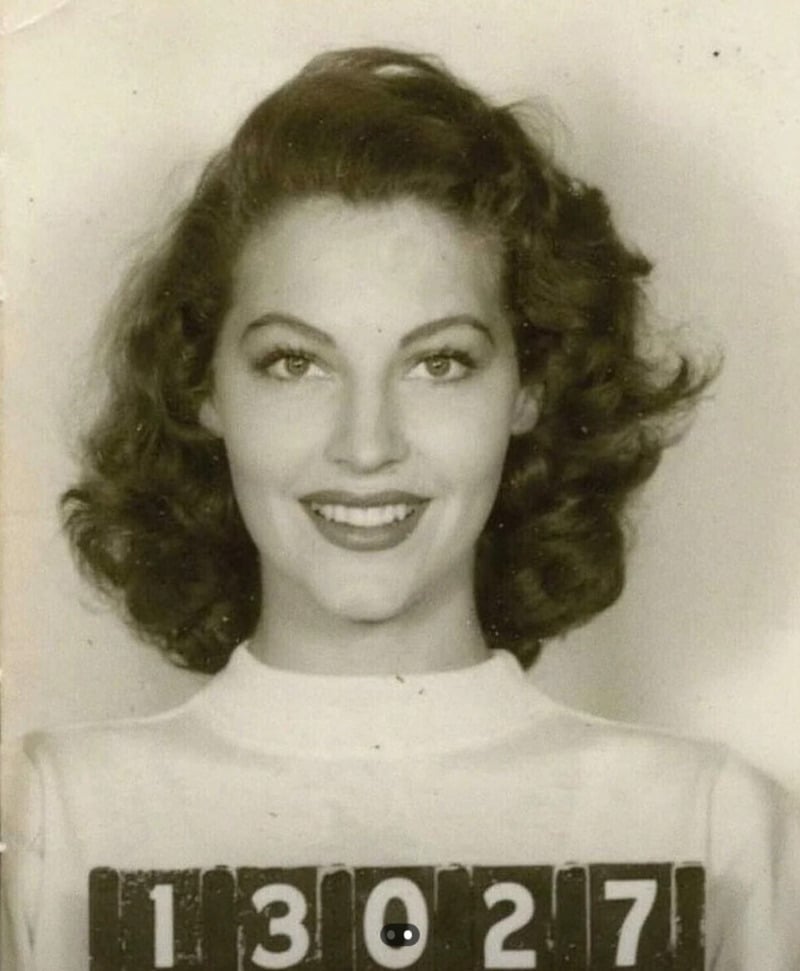 A black and white mugshot of a young woman with wavy hair and a wide smile, wearing a light-colored top. She holds a placard displaying the number 13027.