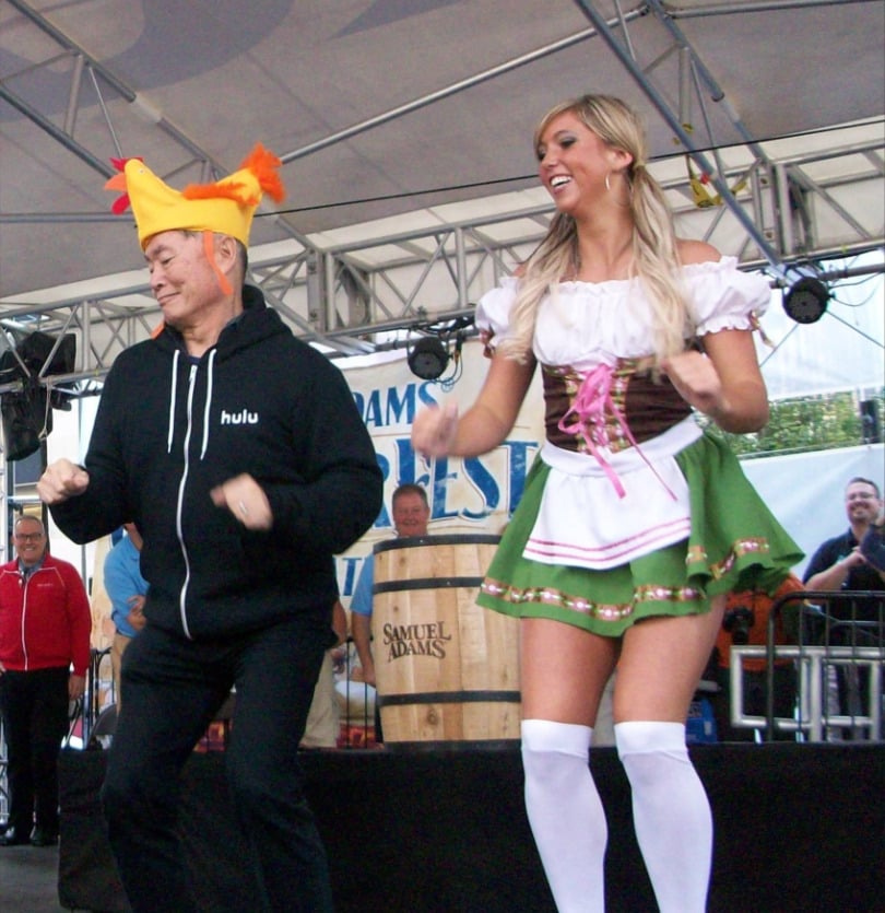 Two people dancing on stage: a man in a yellow chicken hat and "hulu" hoodie, and a woman in a green and white dirndl dress with thigh-high stockings. There are people and a large beer barrel in the background.